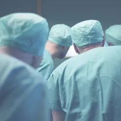 Fire personer i grønt tøy som står utenfor døren til en operasjonsstue og ser inn gjennom vinduet
